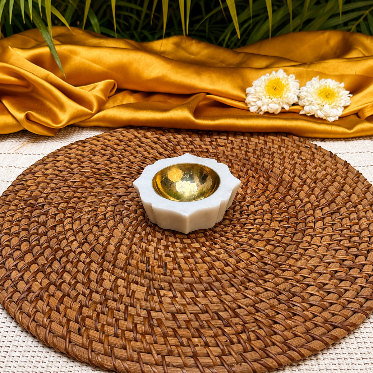 A small white ceramic candle holder with a reflective gold interior, placed on a round woven brown placemat. Gold satin fabric and two white daisy flowers are draped behind it, with green leaves visible at the top edge.