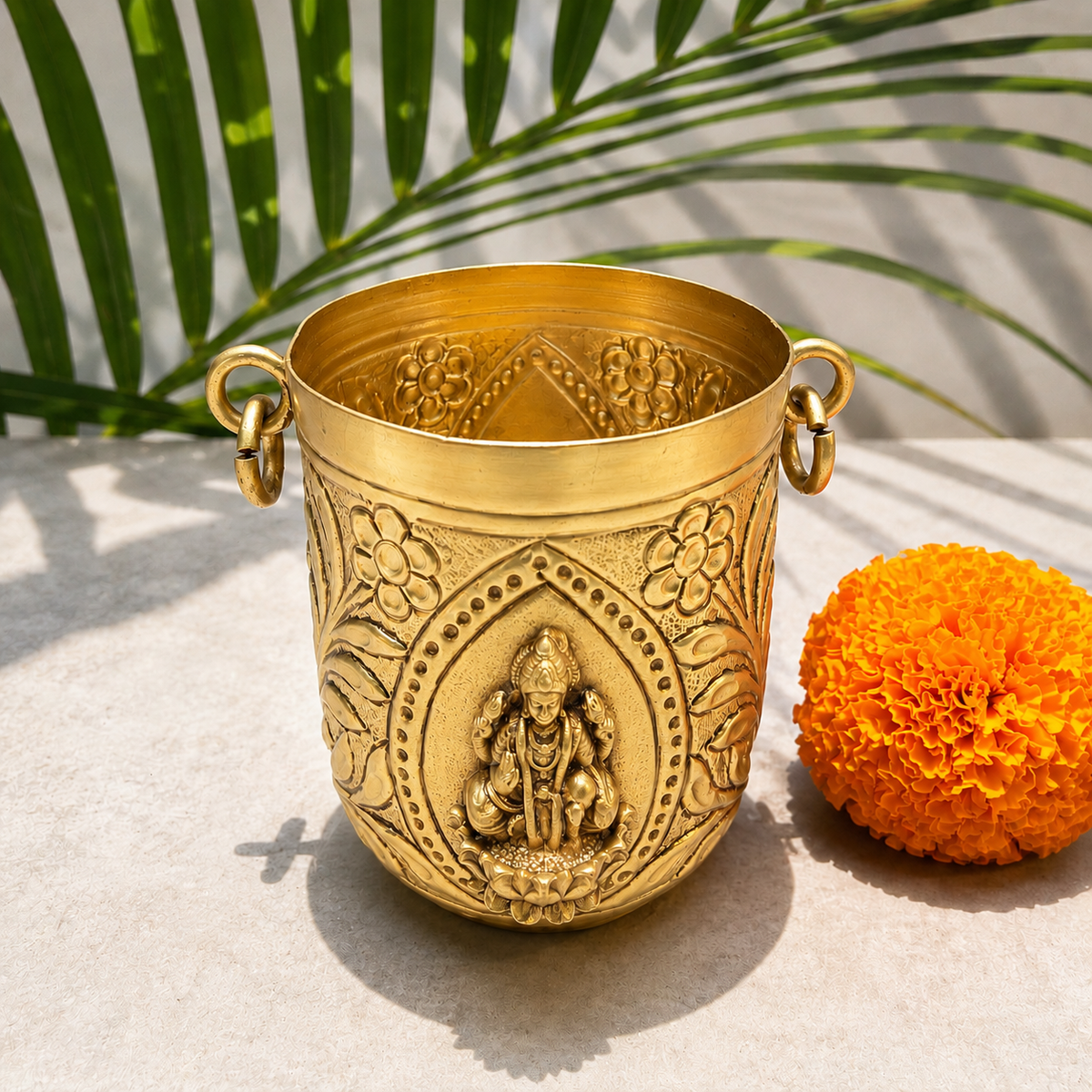 An ornate brass pot with side handles and a carved deity figure on the front, styled on a light stone surface with a marigold flower and green palm leaves in the background.