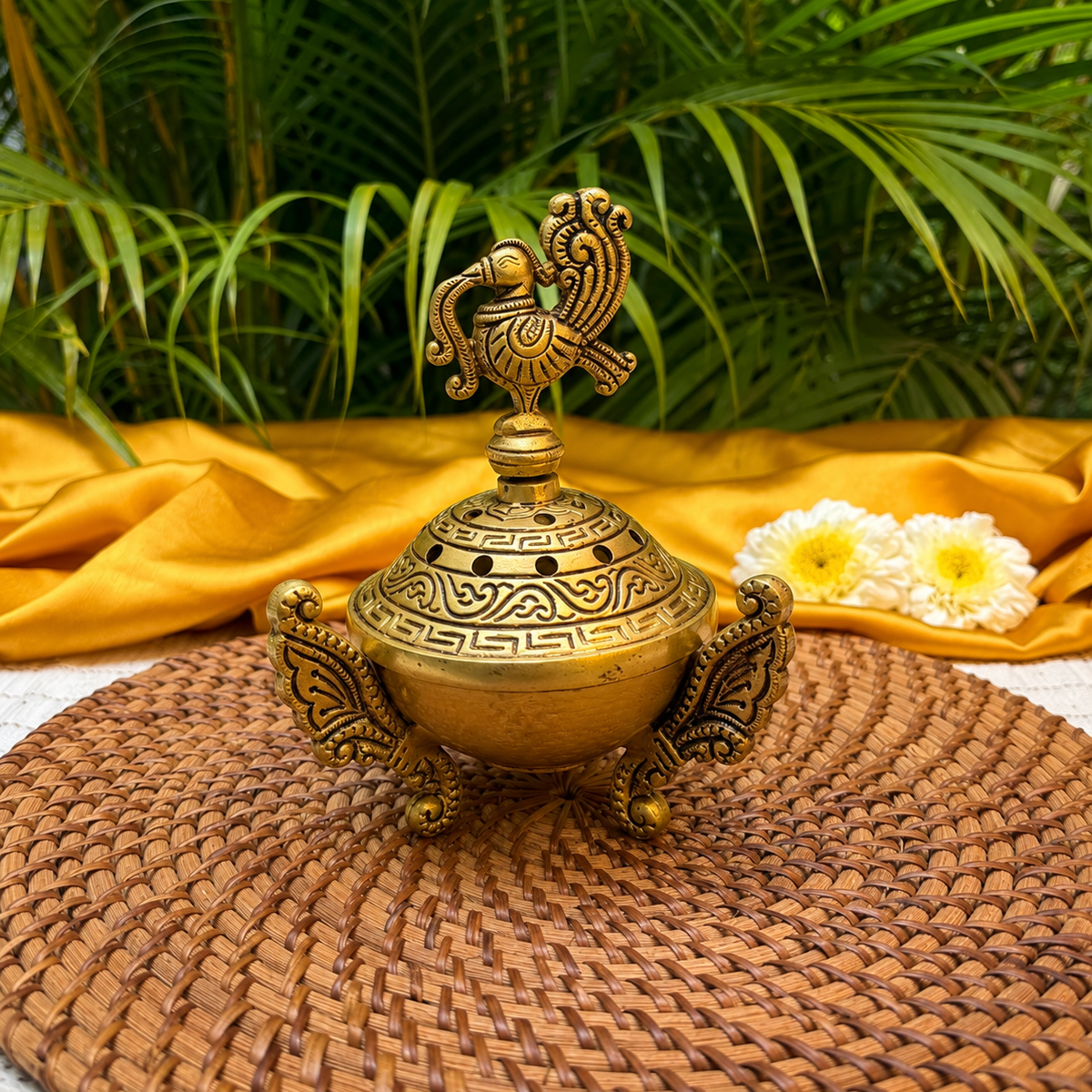 An ornate brass incense burner with a decorative peacock finial on top, set on a woven brown mat with draped yellow fabric and green palm leaves in the background.