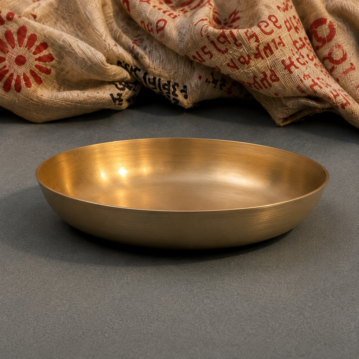 A round, shallow brass-colored metal tray or dish with a brushed metallic finish, set against a dark gray background with a textured burlap sack draped in the upper background.