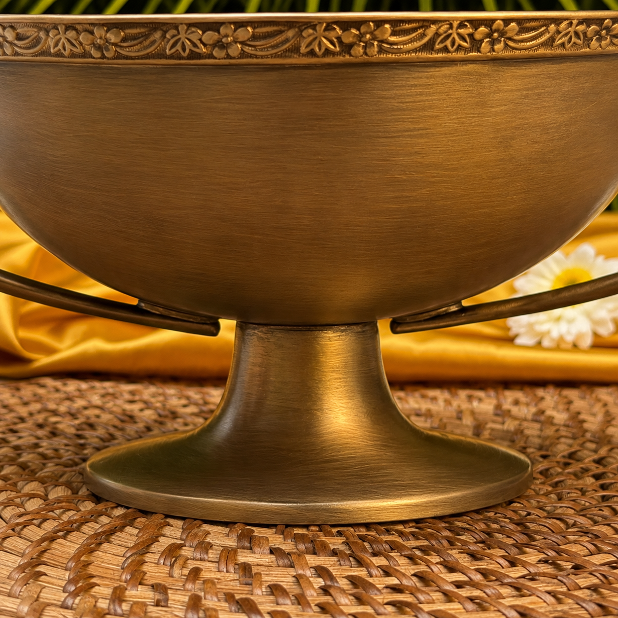 A close-up of an antique bronze pedestal bowl with a decorative floral rim, resting on a woven placemat with yellow fabric and a white flower in the background.
