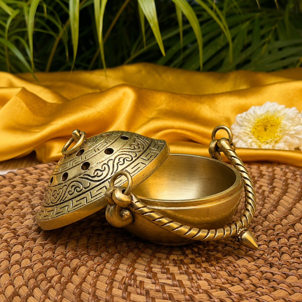 A brass incense burner with an ornate lid and rope handle, styled on a woven mat with gold fabric and green leaves in the background.