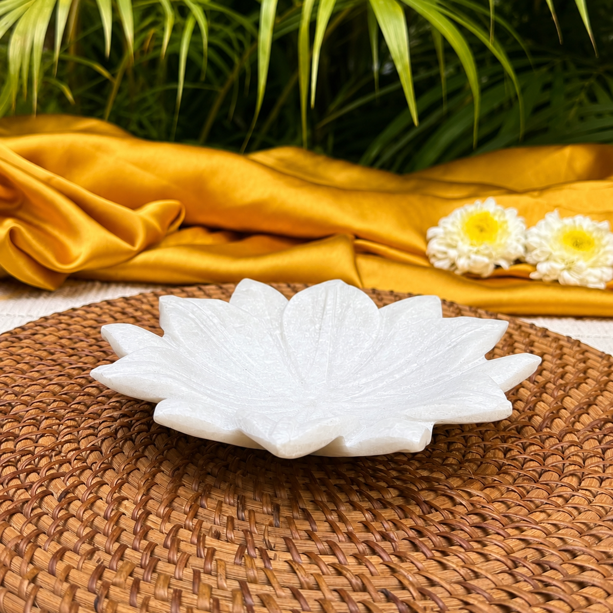 A white lotus-shaped ceramic dish or shallow tray sits on a round woven brown placemat, with draped gold fabric, green leaves, and two white flowers in the background.