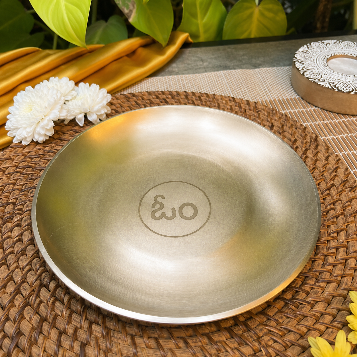 A round brushed-metal plate with a circular engraved symbol in the center, displayed on a woven brown placemat with decorative flowers and green leaves in the background.