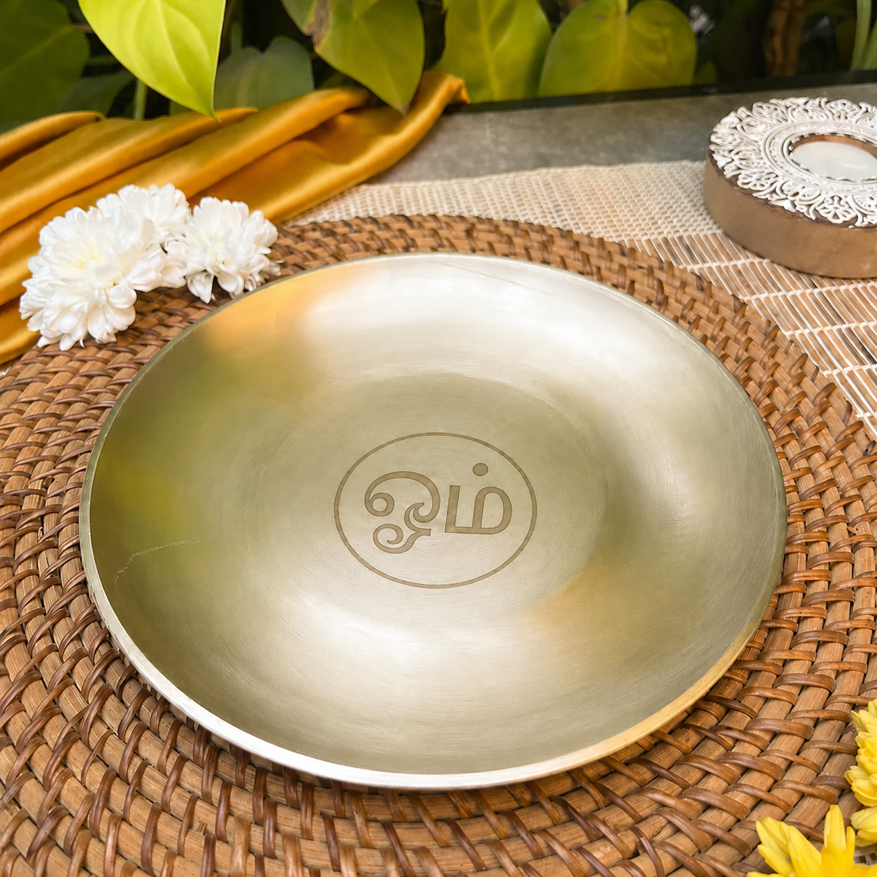 A round metallic gold plate with a custom monogram engraved in the center, displayed on a woven brown placemat with decorative white flowers and green leaves in the background.