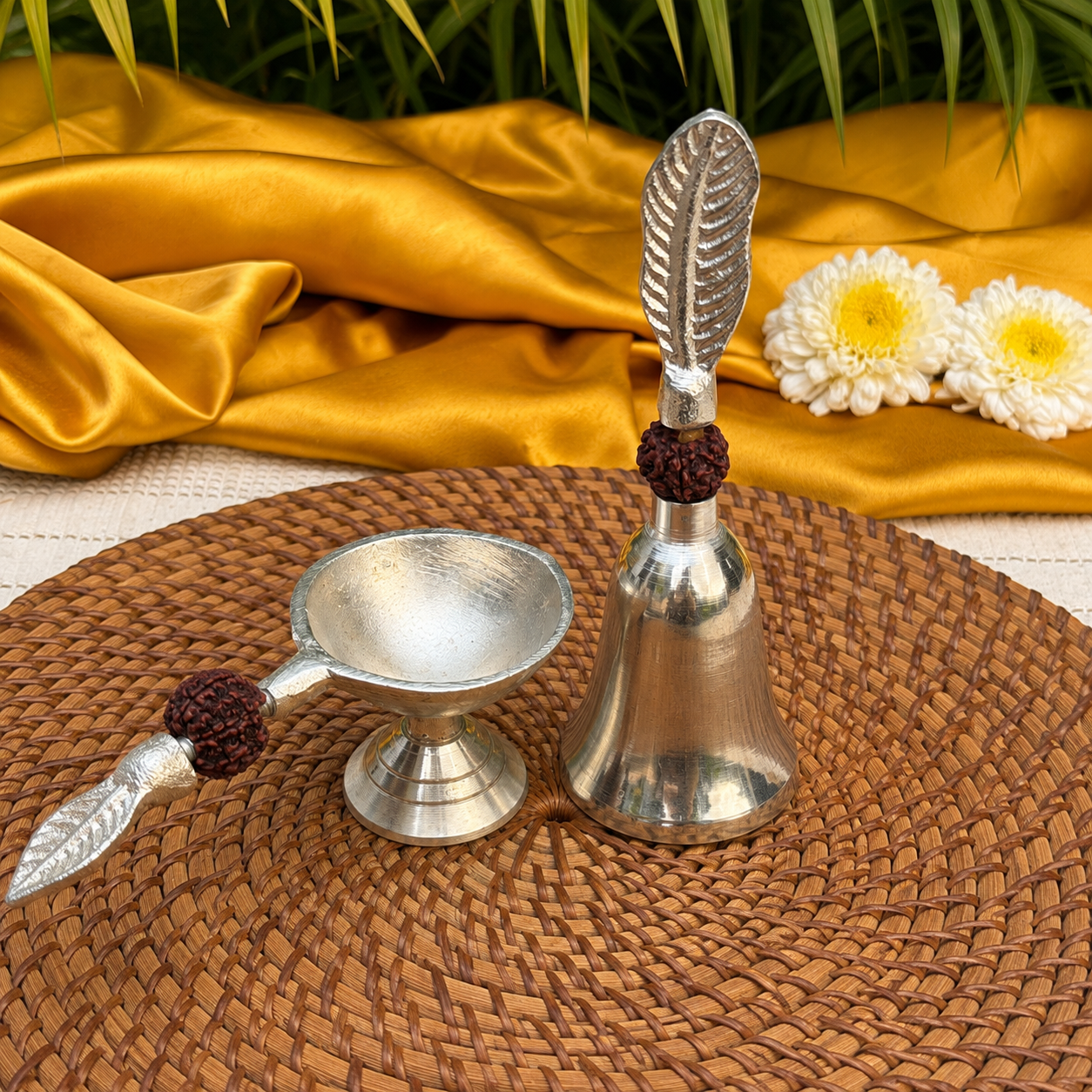 A silver metal diya lamp set with a small bowl and a bell, both featuring leaf-shaped handles and dark red agate beads, arranged on a woven brown placemat with gold fabric and white flowers in the background.