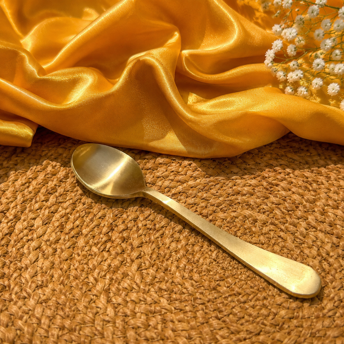 A polished brass spoon resting on a textured brown woven placemat, with draped golden-yellow satin fabric and small white flowers in the background.