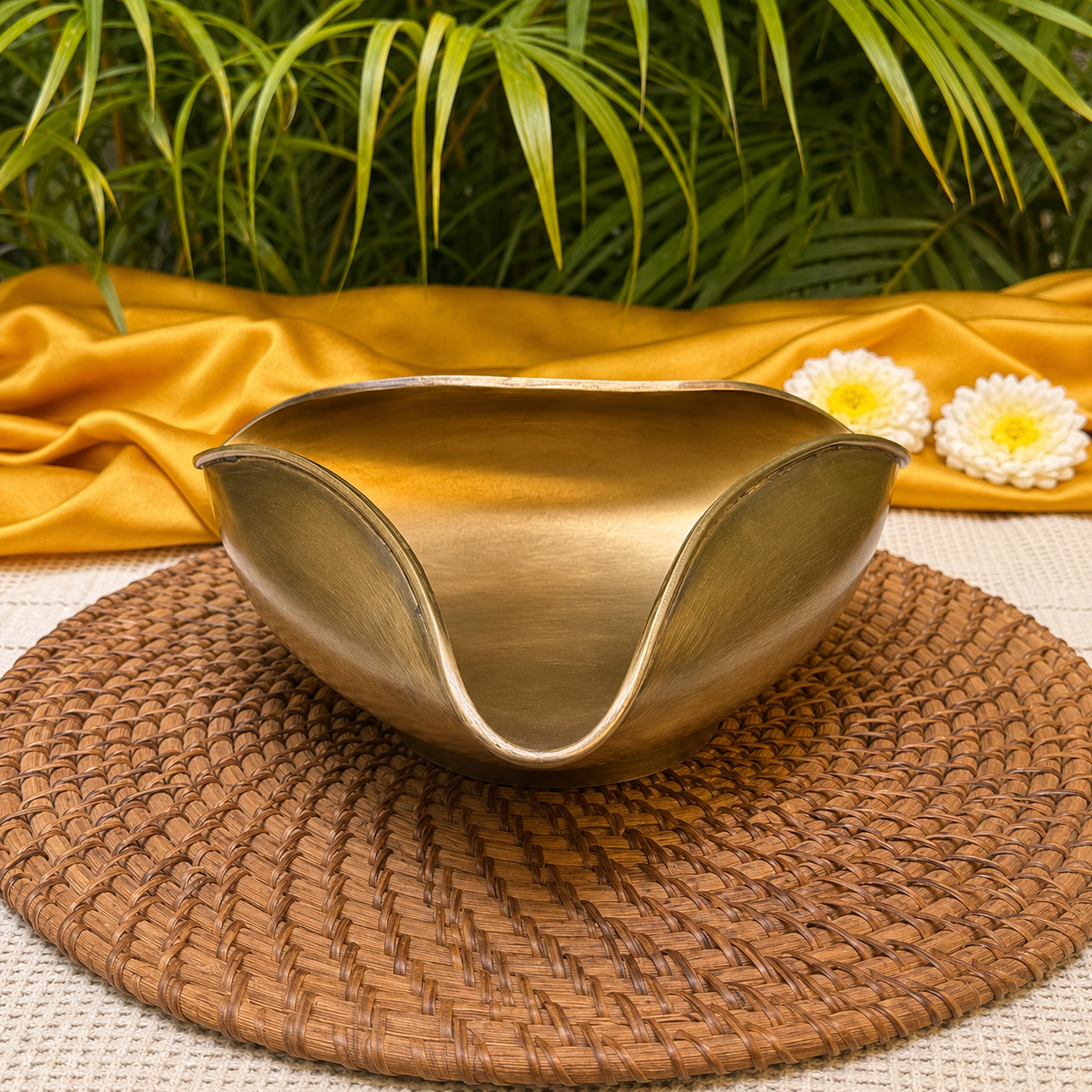 A hammered brass serving bowl with a unique wavy rim, displayed on a round woven placemat with a draped mustard yellow fabric and green foliage in the background.
