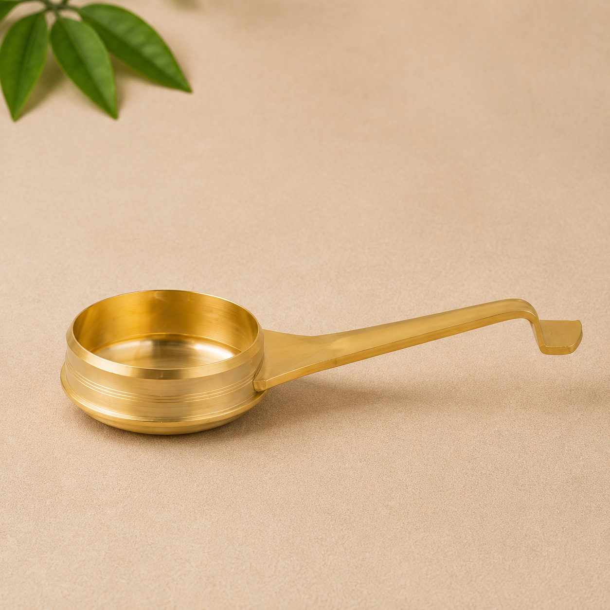 A brass ladle with a long handle and a looped end, shown on a textured beige background with a small green leaf in the top-left corner.