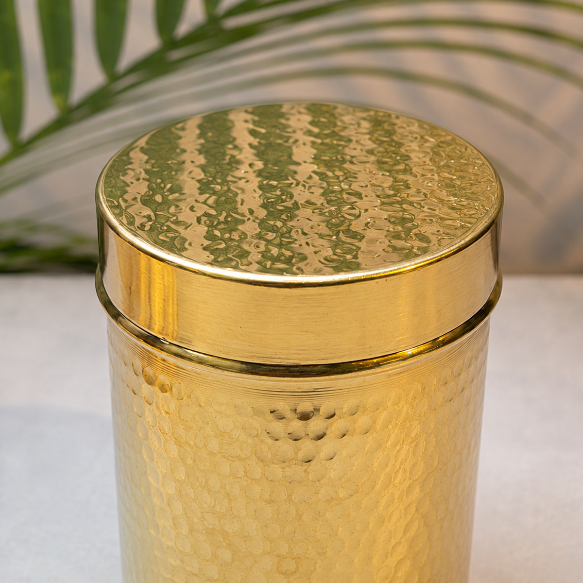 A close-up of a cylindrical gold-toned container with a hammered metallic finish and a matching lid, shown against a softly blurred background with a green leaf.