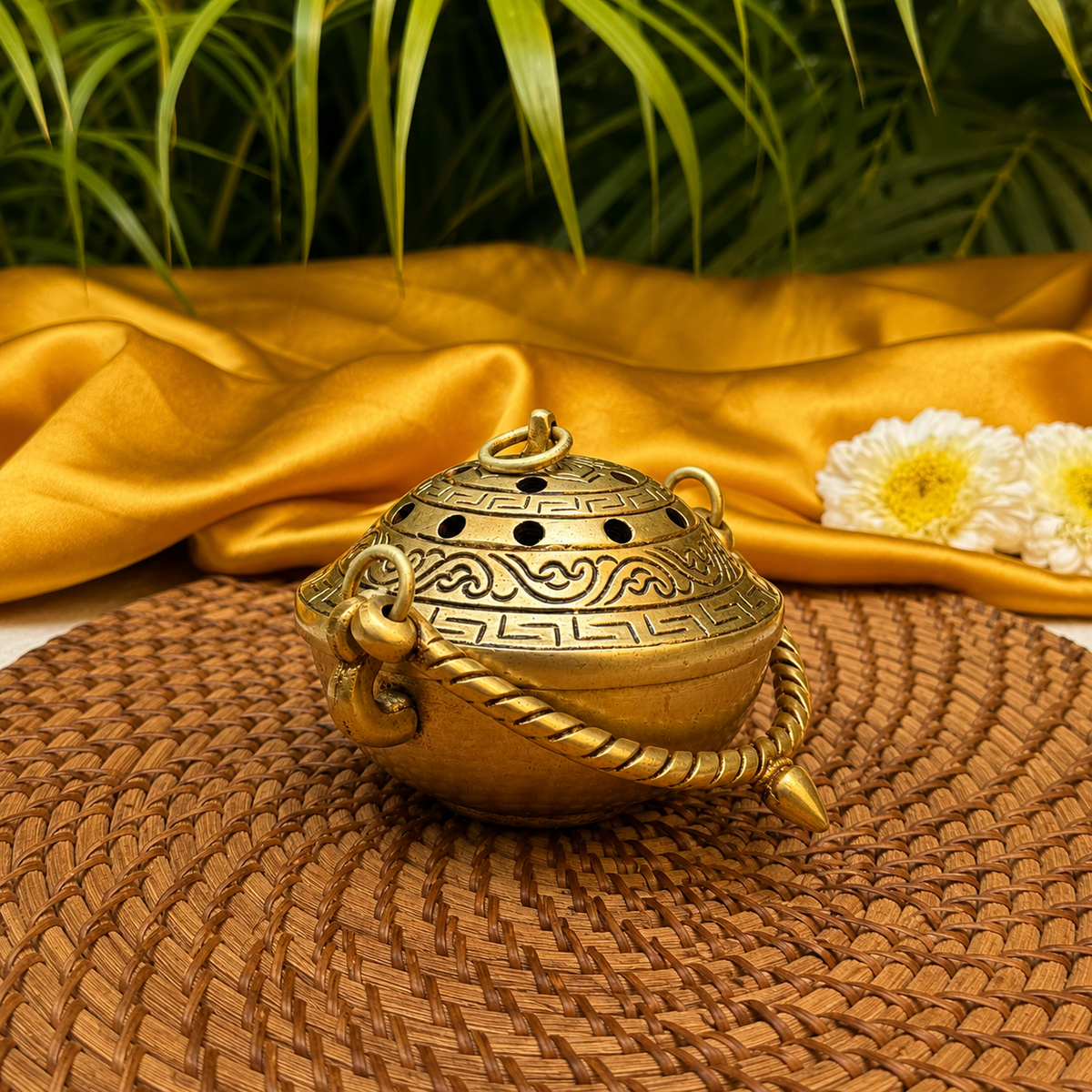 An ornate brass incense burner with engraved patterns and a loop handle, placed on a woven brown mat with draped gold fabric, green leaves, and white flowers in the background.