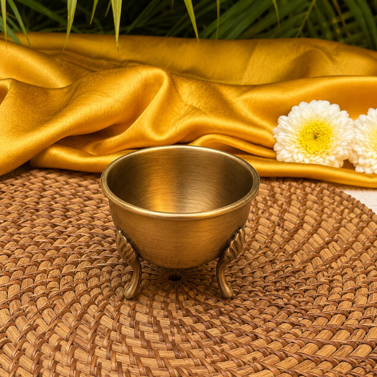 A small three-legged brass bowl sits on a woven round mat, with draped gold fabric and white flowers in the background.