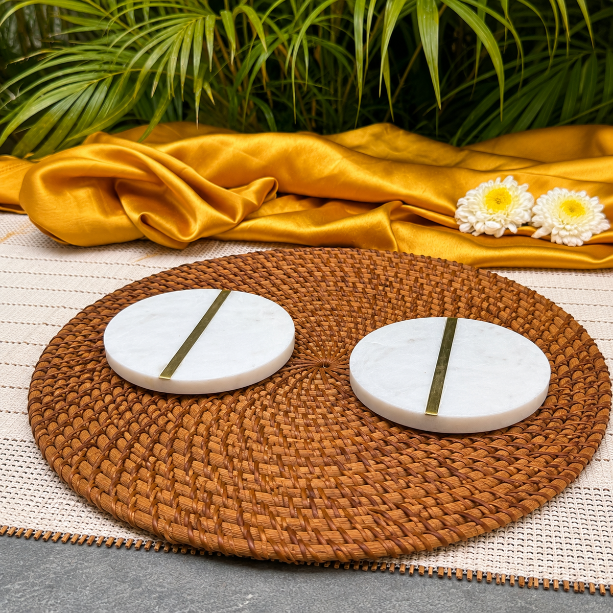 Two round white marble coasters with a central gold metallic stripe, displayed on a round brown woven placemat with a gold satin cloth and green leaves in the background.