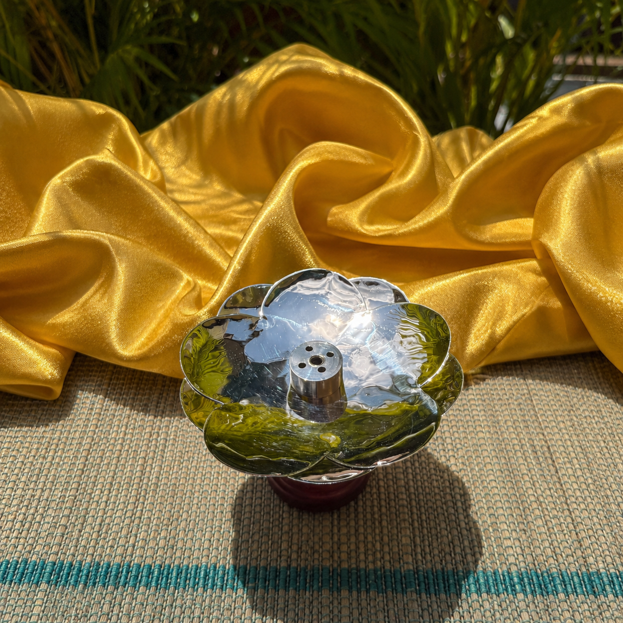 A clear glass lotus-shaped candle holder with green leaf-patterned inlay and a silver-toned metal top, set on a woven mat with draped yellow silk fabric in the background.