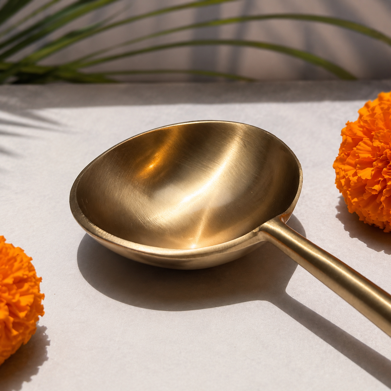 A brushed brass-colored metal serving spoon or ladle with a long handle, styled on a light surface with orange marigold flowers and soft shadows.