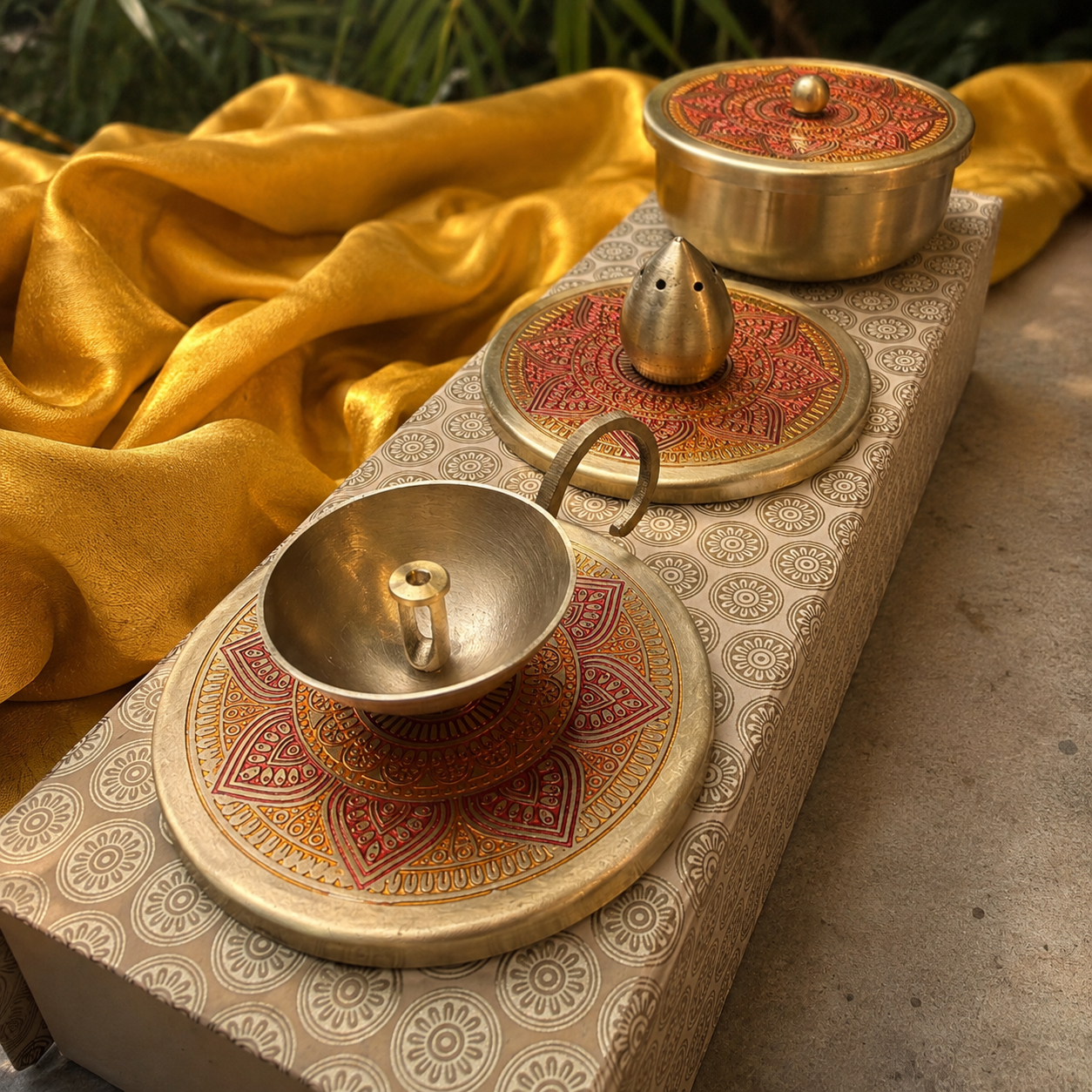Three brass temple idols or decorative idols with ornate red and gold patterned lids, arranged on a patterned rectangular box with draped yellow fabric in the background.