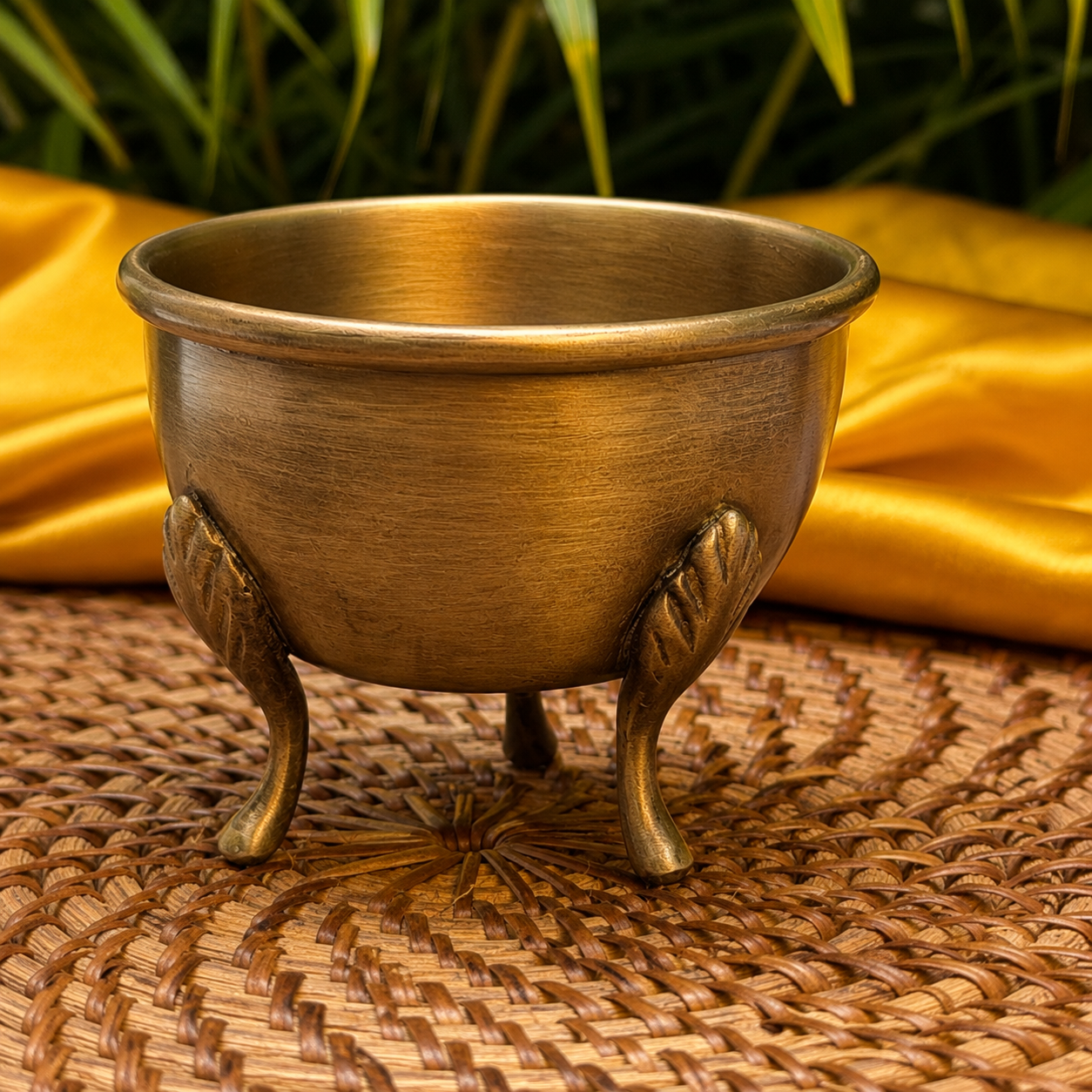 A small brass-colored metal bowl with three decorative legs, shown on a woven mat with gold fabric and green leaves in the background.