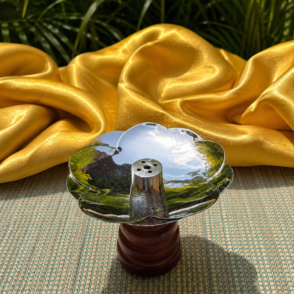A polished silver-toned metal flower-shaped object on a dark brown wooden stand, set on a woven mat with draped golden-yellow fabric and green foliage in the background.