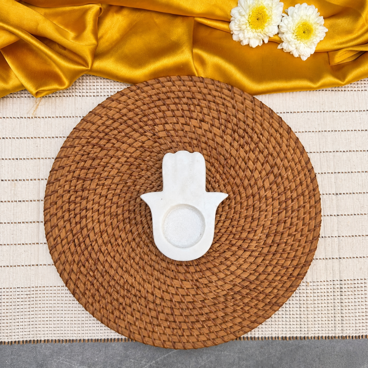 A round woven brown placemat on a cream textured table runner, styled with a white ceramic Hamsa hand dish in the center and draped gold satin fabric with white flowers at the top.