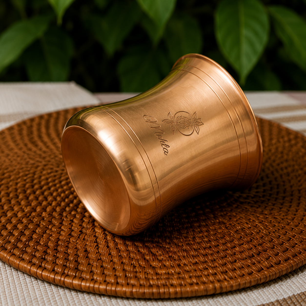 A copper-colored metal tumbler with engraved script and a floral motif, resting on a round woven brown placemat with a blurred green leafy background.