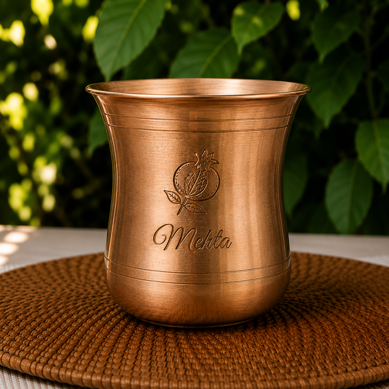 A copper-colored metal mug with an engraved pomegranate logo and the name 'Mehta' on the front, sitting on a woven brown placemat with a blurred green leafy background.