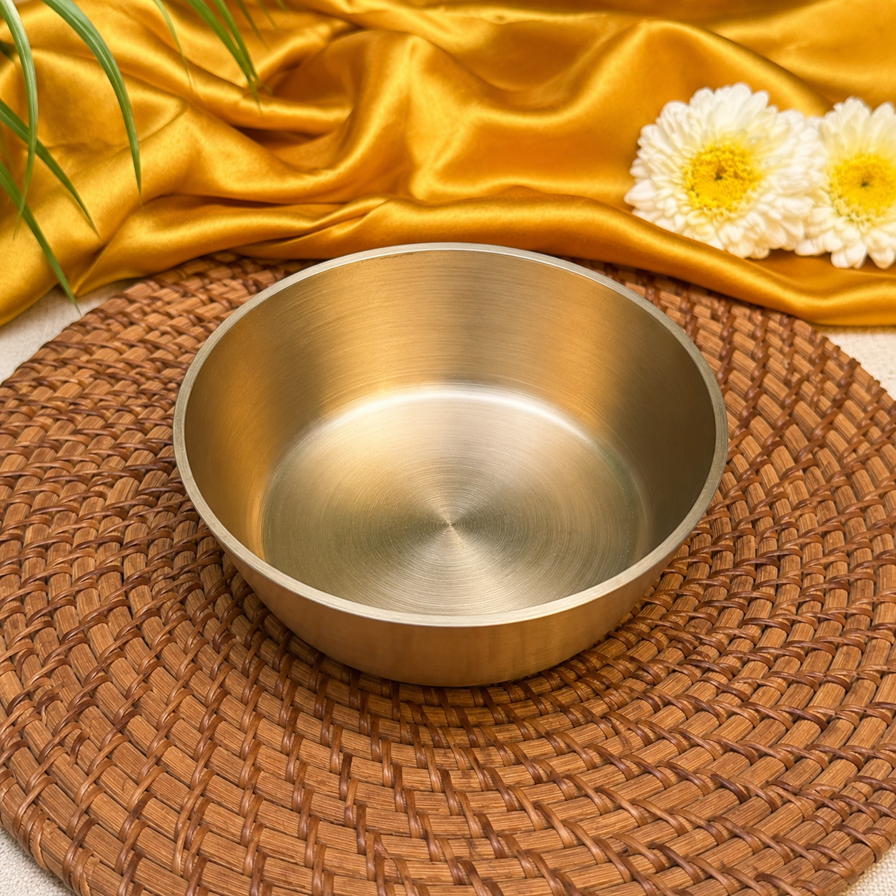 A brushed metal bowl with a warm gold finish, resting on a brown woven placemat with draped gold fabric and white flowers in the background.
