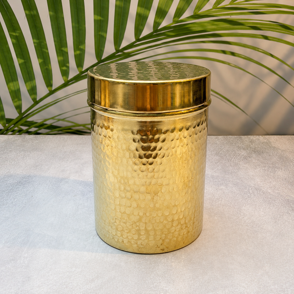 A cylindrical hammered brass container with a matching lid, set on a light textured surface with a green palm leaf in the background.
