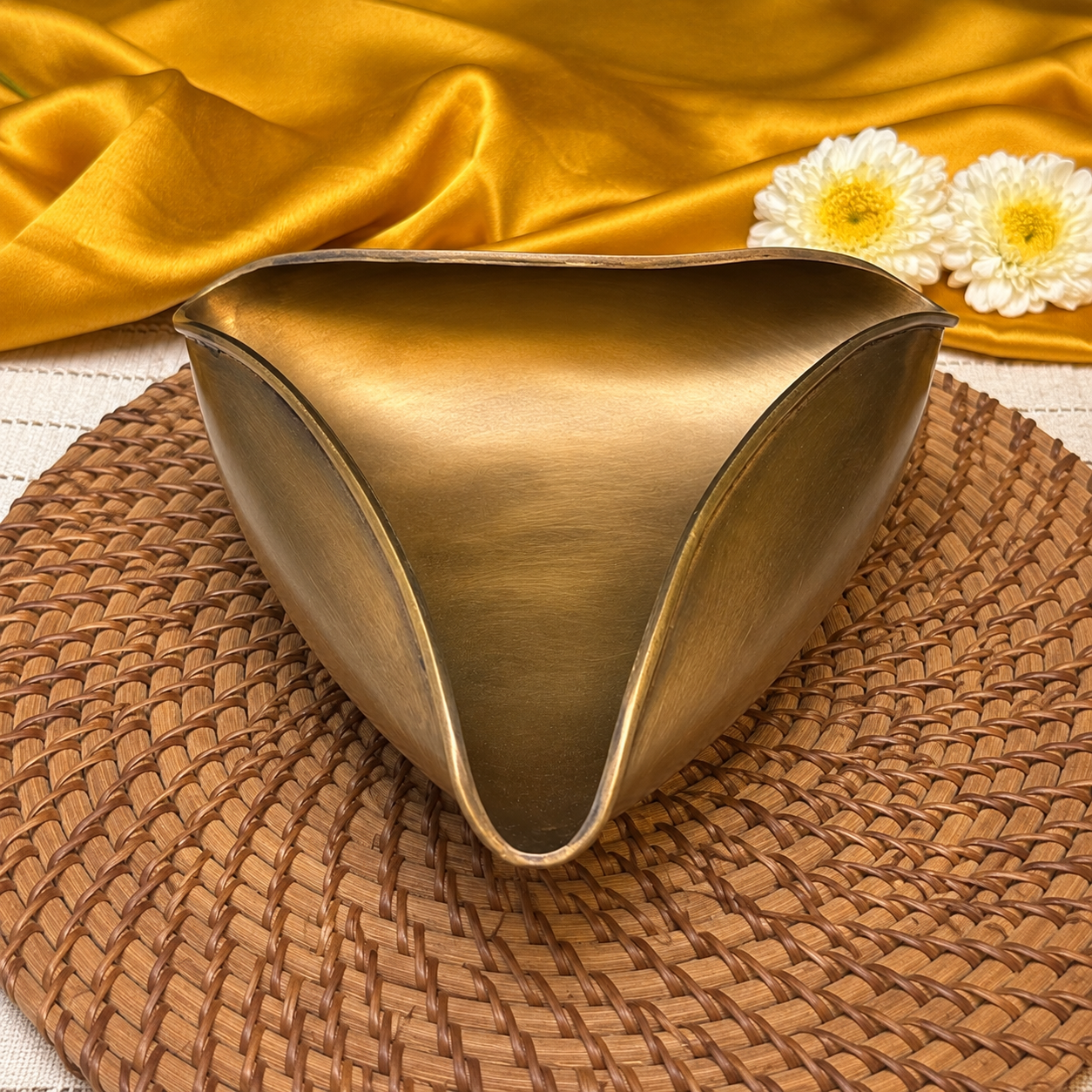 A metallic gold bowl with a curved, organic shape, resting on a brown woven placemat with a draped gold fabric and white daisy flowers in the background.