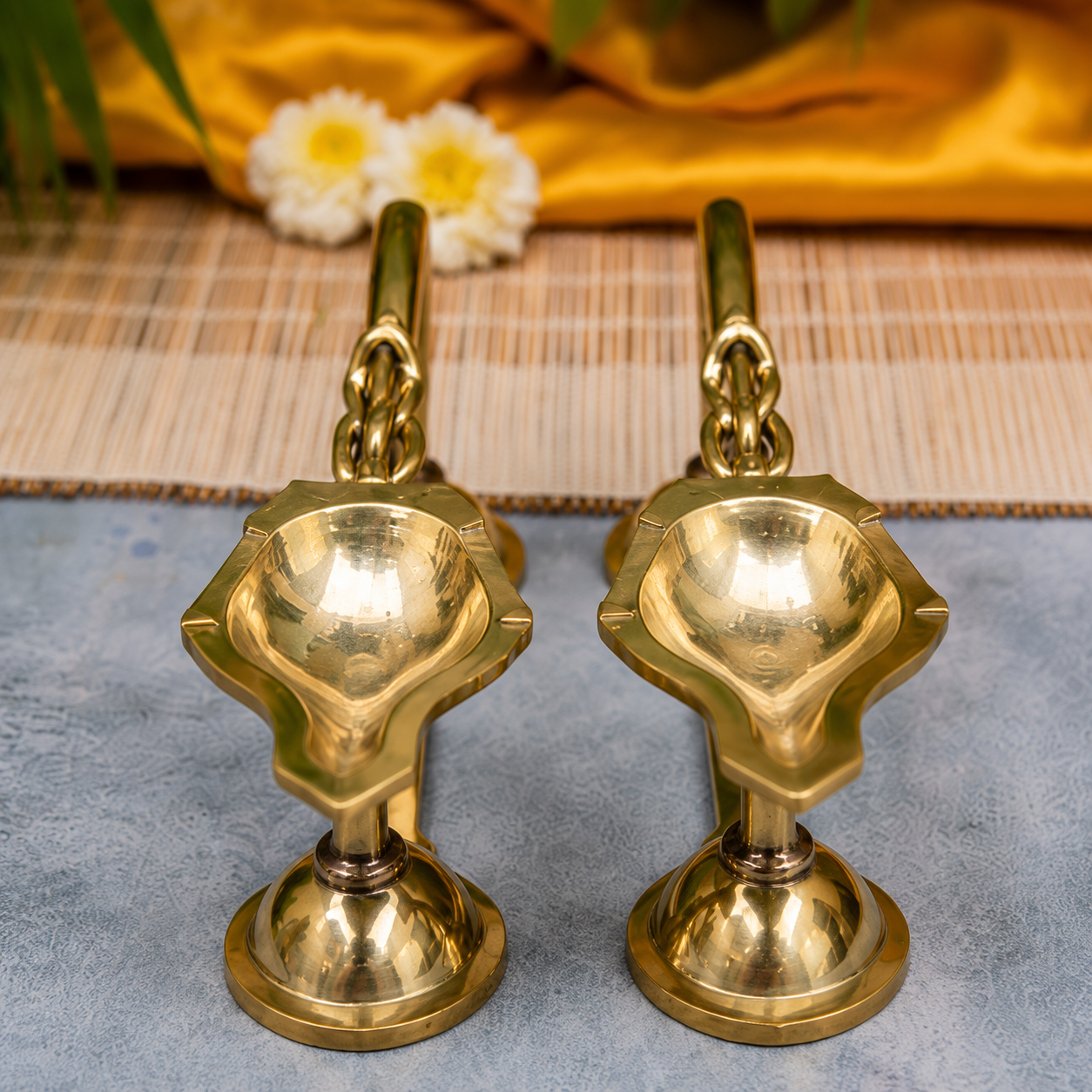Two polished brass oil lamps with decorative floral-shaped bases, displayed side by side on a textured gray surface with a woven mat and yellow fabric in the background.