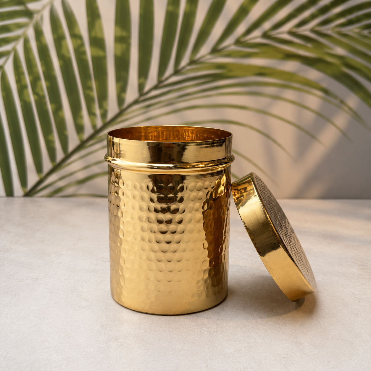 A hammered gold metal tea or spice container with its lid leaning against it, set on a light surface with a green palm leaf backdrop.