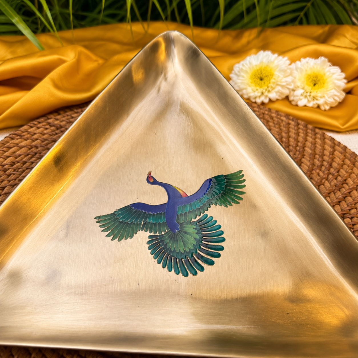 A triangular brass tray featuring an inlaid colorful phoenix bird design in shades of blue, green, and gold, styled on a woven mat with yellow fabric and white flowers in the background.