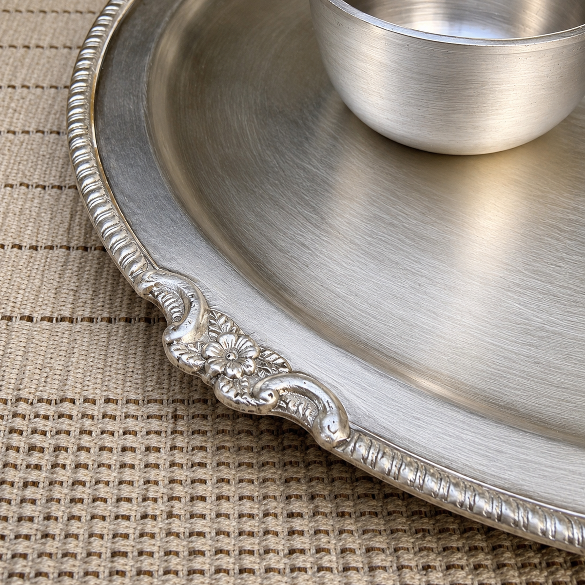 A close-up of a brushed silver metal serving platter with an ornate decorative handle, resting on a woven beige placemat with a matching small metal cup partially visible in the upper right.
