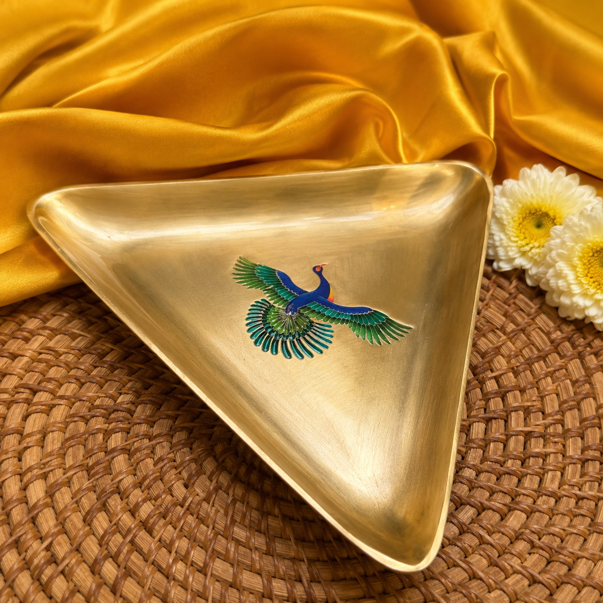A triangular brass tray with a polished gold finish, featuring an inlaid peacock design in vibrant blue, green, and orange. The tray is styled on a brown woven placemat with draped golden-yellow satin fabric and white daisies in the background.