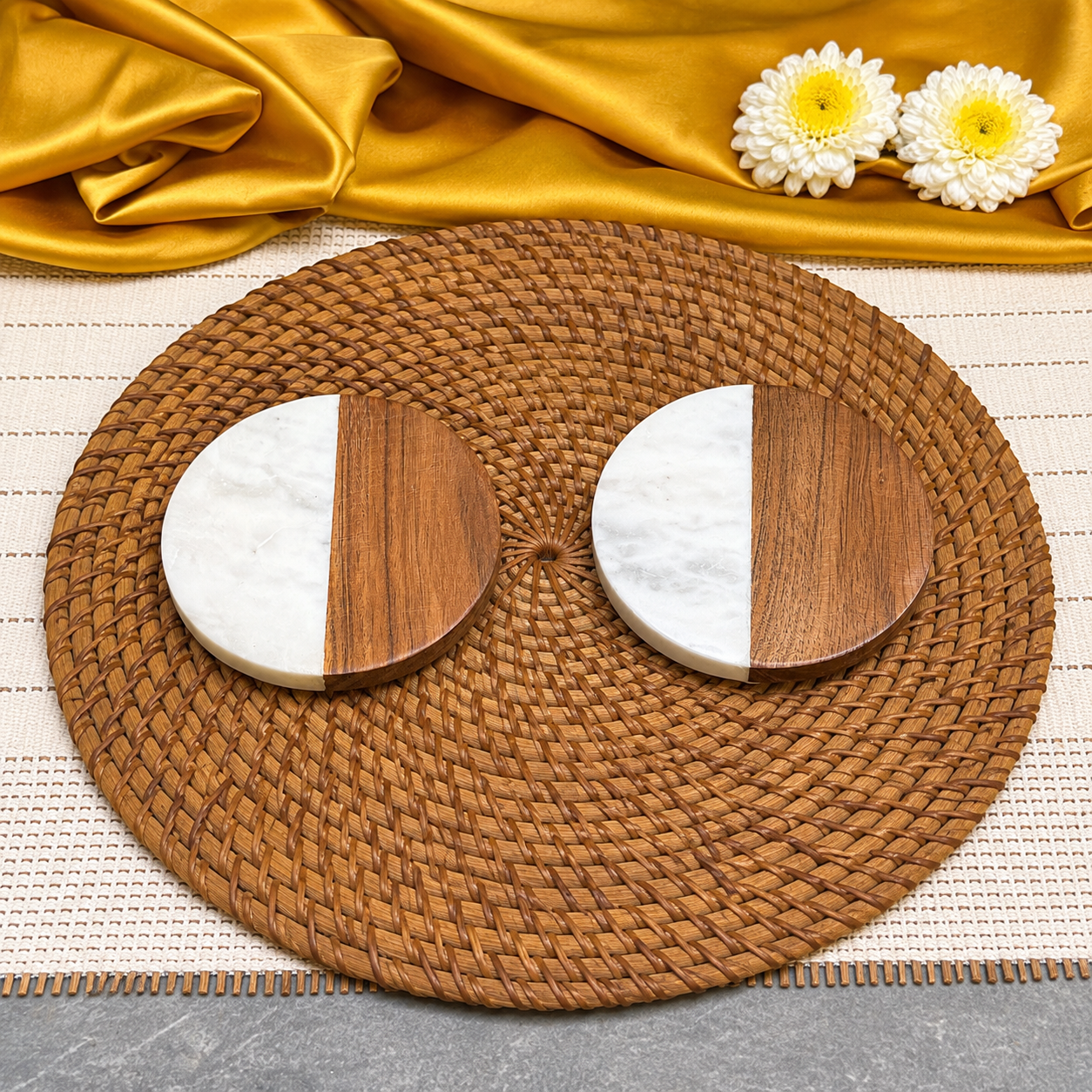 Two round coasters with a split white marble and dark wood design, arranged on a round woven brown placemat with gold fabric and white flowers in the background.