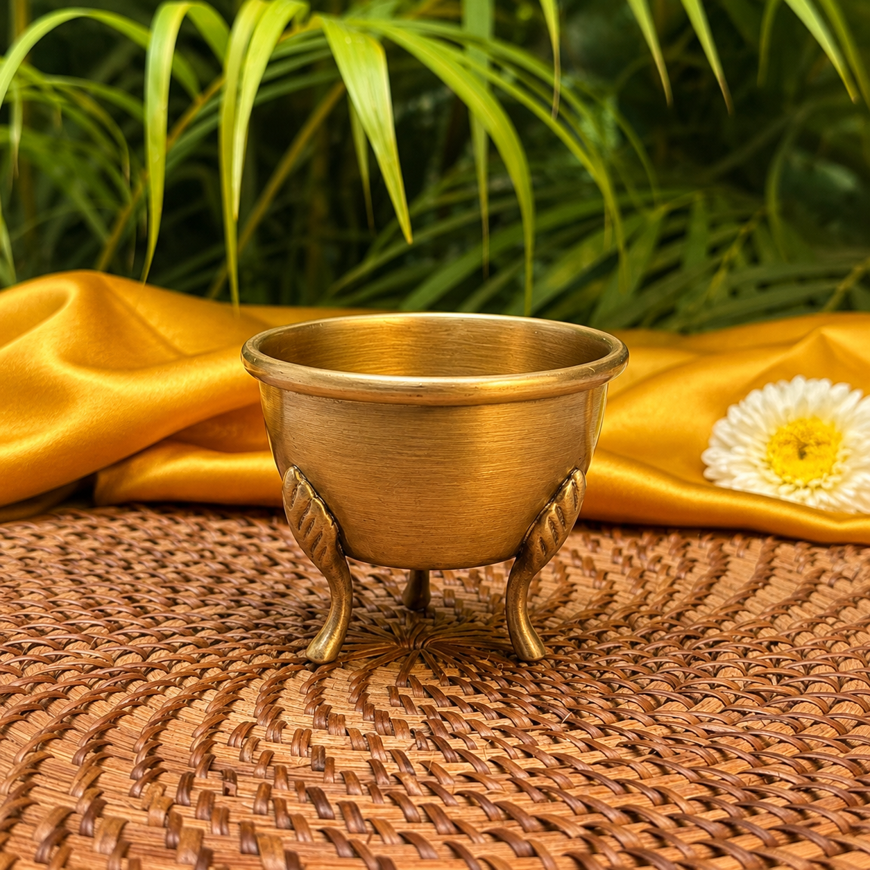 A small brass-colored metal cup with three decorative legs sits on a woven brown placemat, with gold satin fabric and green leaves in the background.