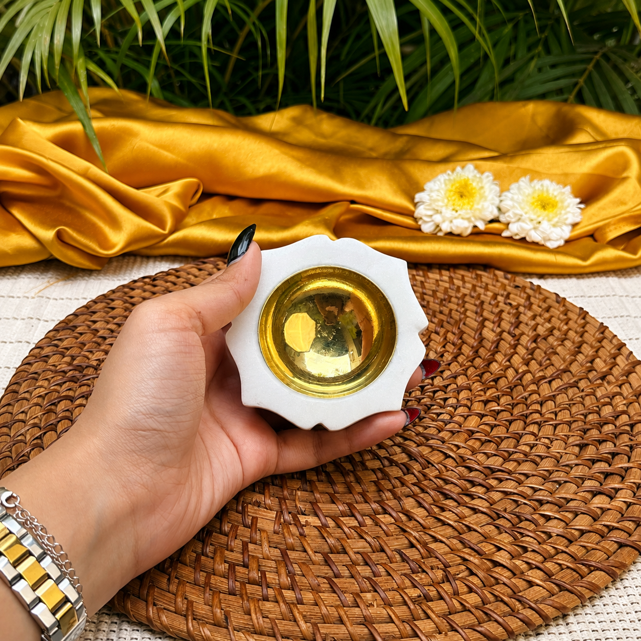 A hand holds a small white ceramic cup with a gold interior, set on a woven brown placemat with gold fabric, white flowers, and green leaves in the background.