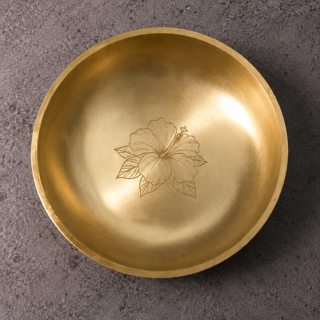 A round brass bowl with a hand-painted hibiscus flower design in the center, photographed from directly above on a textured gray surface.