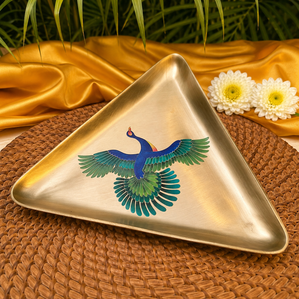A triangular brass tray featuring a colorful hand-painted peacock, styled on a brown woven placemat with gold satin fabric and white flowers in the background.