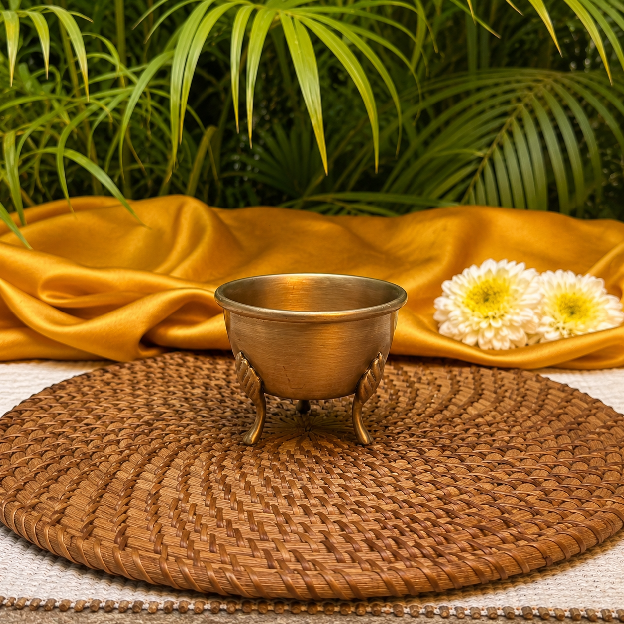 A small brass pedestal bowl with three legs sits centered on a round woven placemat, styled with draped mustard-yellow fabric, white and yellow flowers, and green palm leaves in the background.