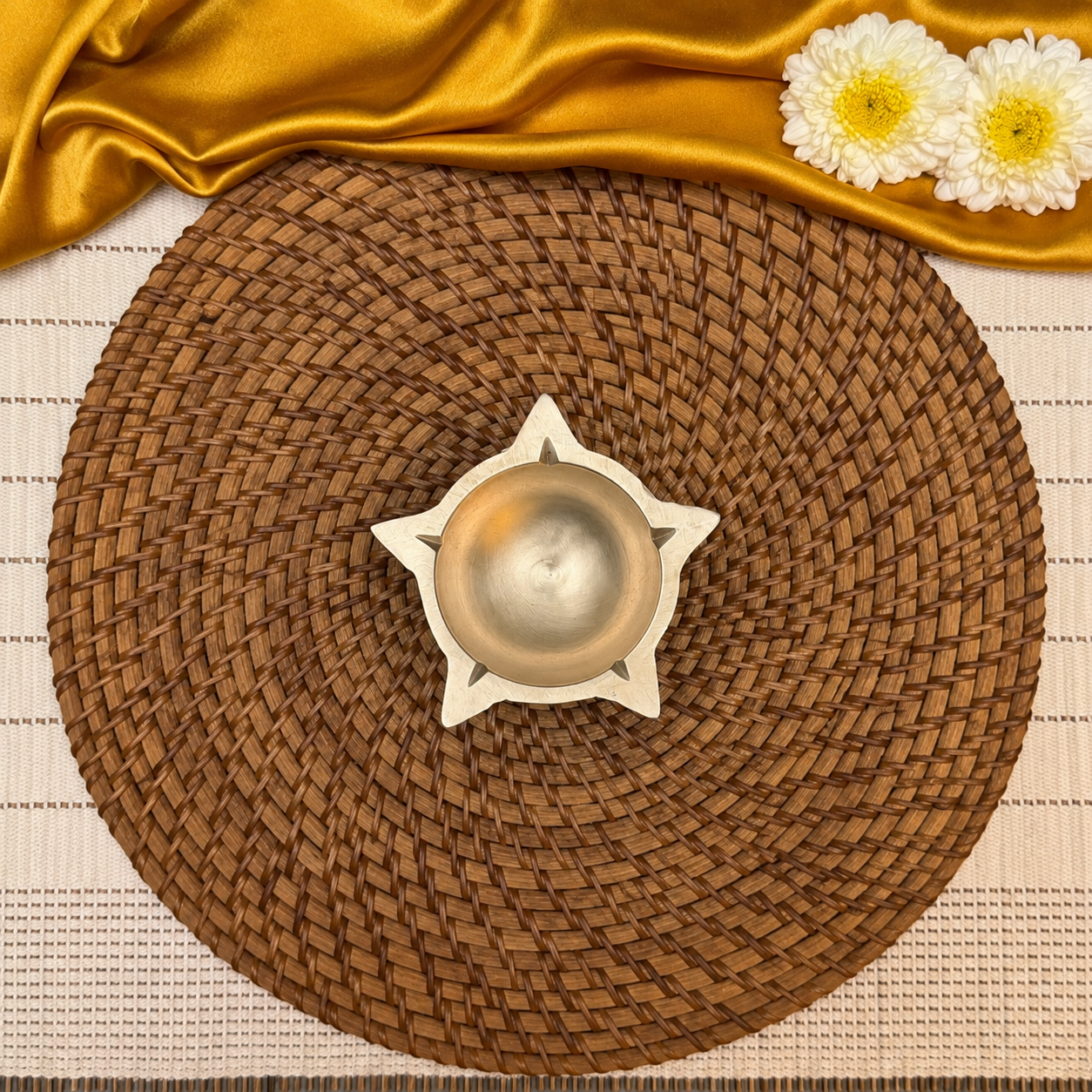 A round brown woven placemat with a star-shaped white holder in the center containing a metallic gold bowl, styled with a gold satin cloth and white flowers on a striped beige and white table runner.
