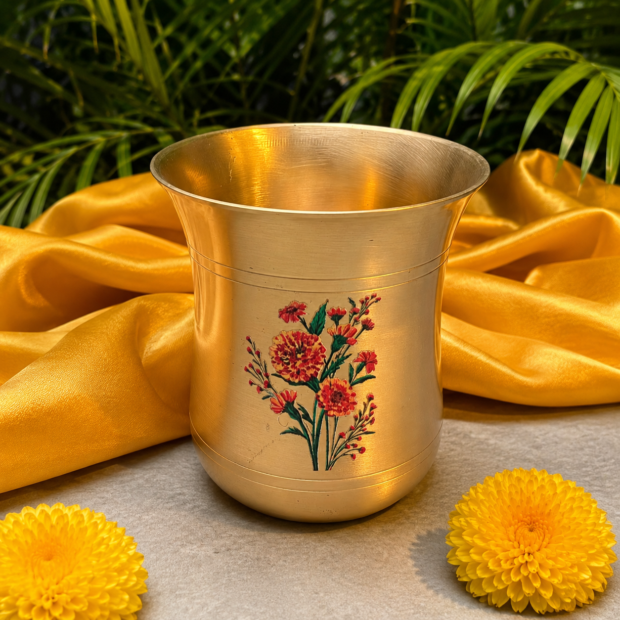 A polished brass cup decorated with a red and green floral illustration, set against a draped yellow silk fabric with green leaves in the background and yellow flowers in the foreground.