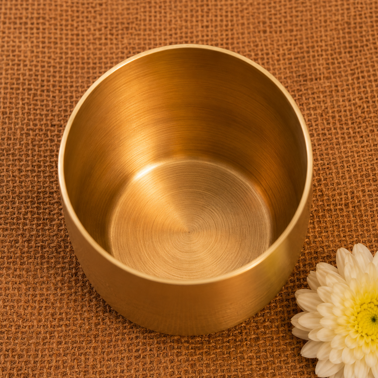 A small round brushed metal cup or bowl in a warm gold/brass tone, photographed from above on a brown woven fabric surface with a white flower partially visible at the lower right.