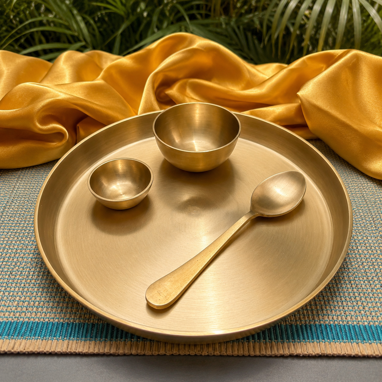 A set of brushed brass tableware, including a large round platter, a small bowl, a spoon, and another miniature bowl, arranged on a woven blue and beige placemat with draped gold fabric and green foliage in the background.