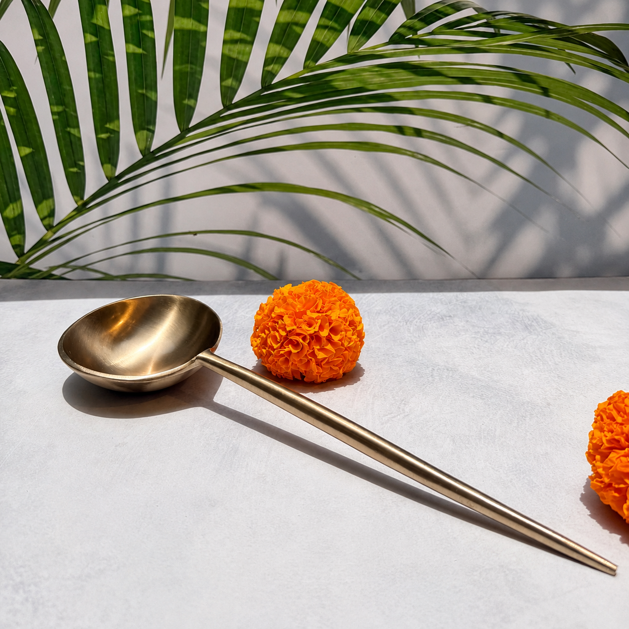 A long-handled brass spoon with a polished metallic finish, styled on a light gray textured surface with orange marigold flowers and a green palm leaf in the background.