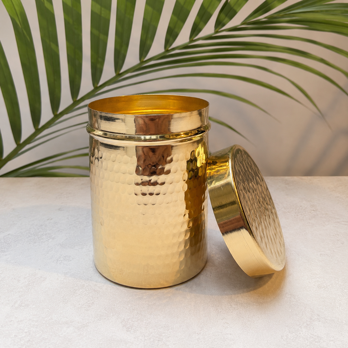 A hammered brass cylindrical container with its lid leaning against it, set on a light stone surface with a green palm leaf backdrop.