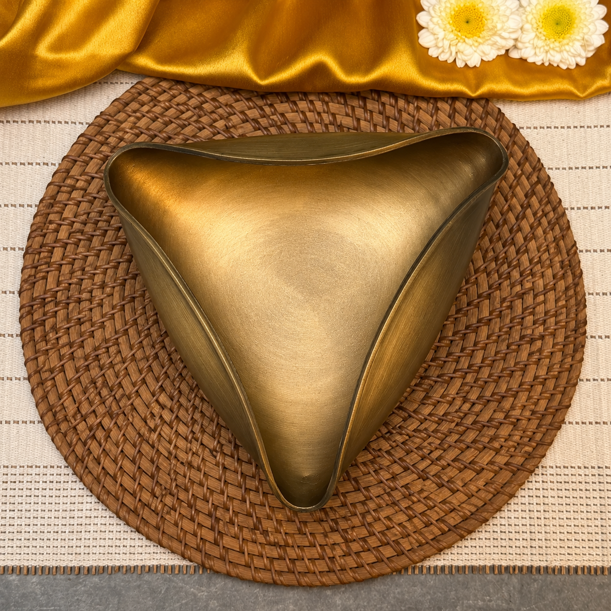 A triangular metallic bowl with a brushed gold finish, styled on a round woven brown placemat with a draped gold fabric and white daisies at the top.