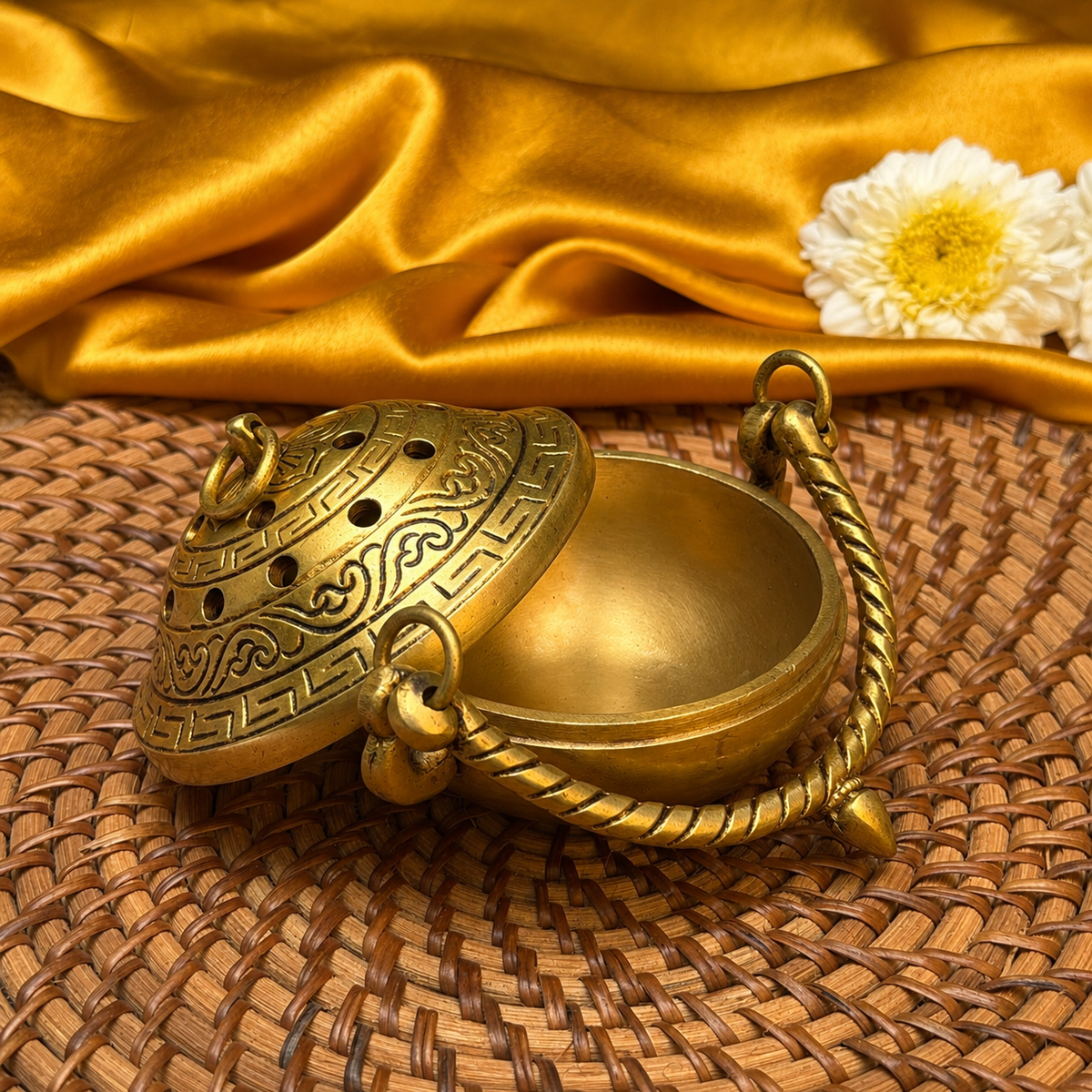 A brass incense burner with ornate engraved patterns and a twisted handle, shown open on a woven mat with draped gold satin fabric and a white flower in the background.