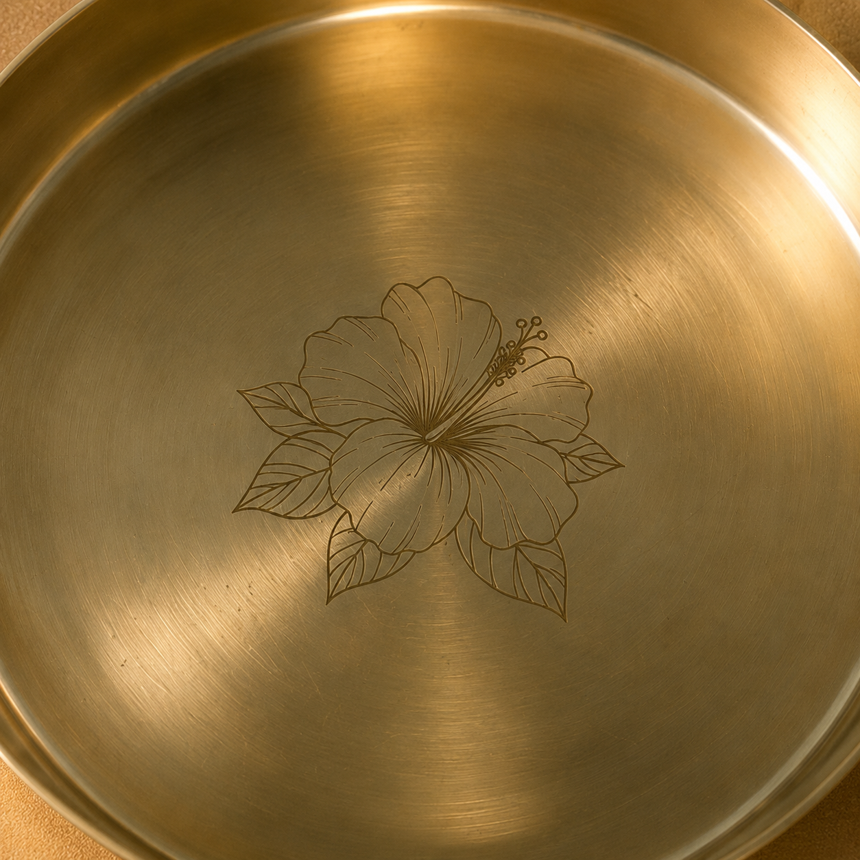 Close-up of the inside of a round metallic pan or tray with a brushed gold finish and an engraved hibiscus flower design in the center.