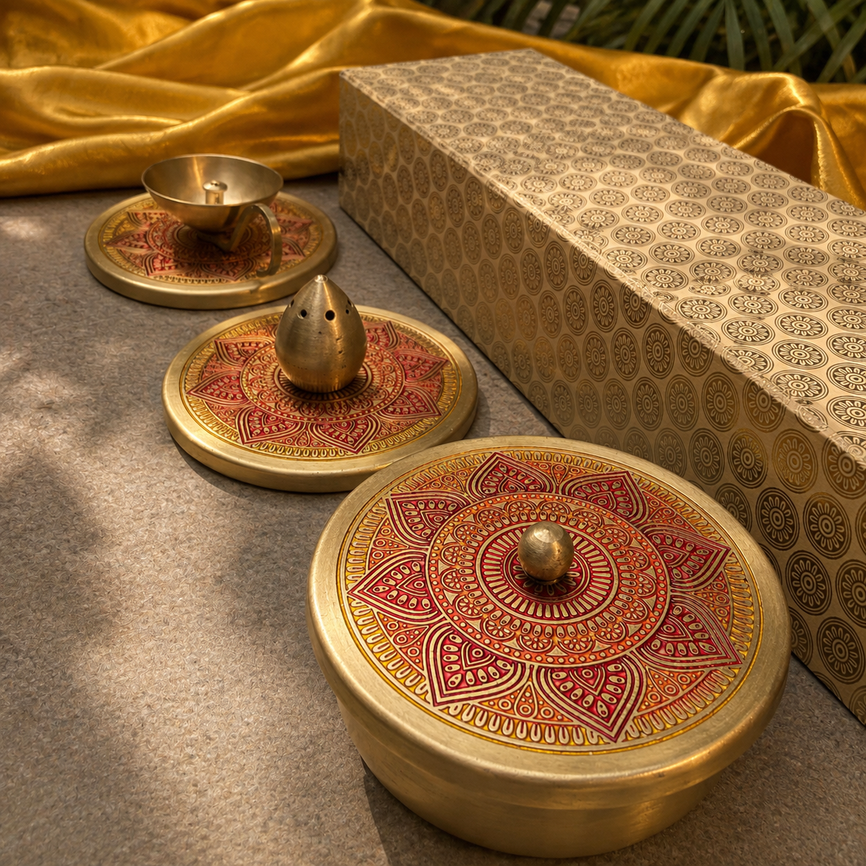 Three round brass keepsakes with ornate red and gold mandala-patterned lids are arranged on a textured gray surface next to a patterned gift box and draped gold fabric.