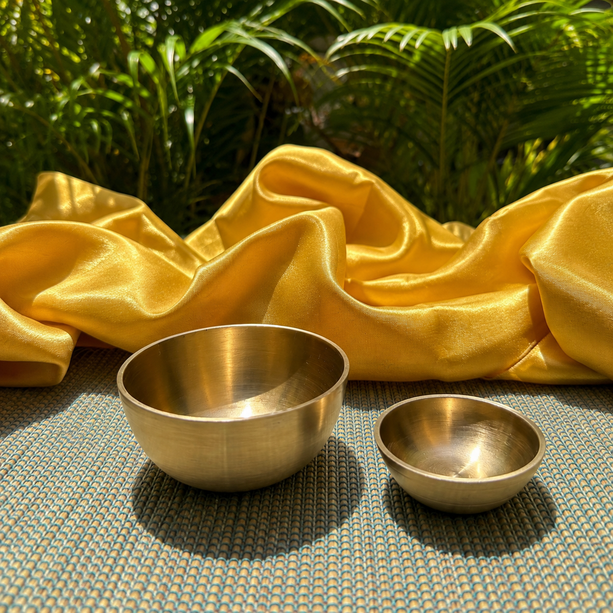 Two brass singing bowls, one large and one small, placed on a woven mat with a draped gold fabric backdrop and green foliage in the background.